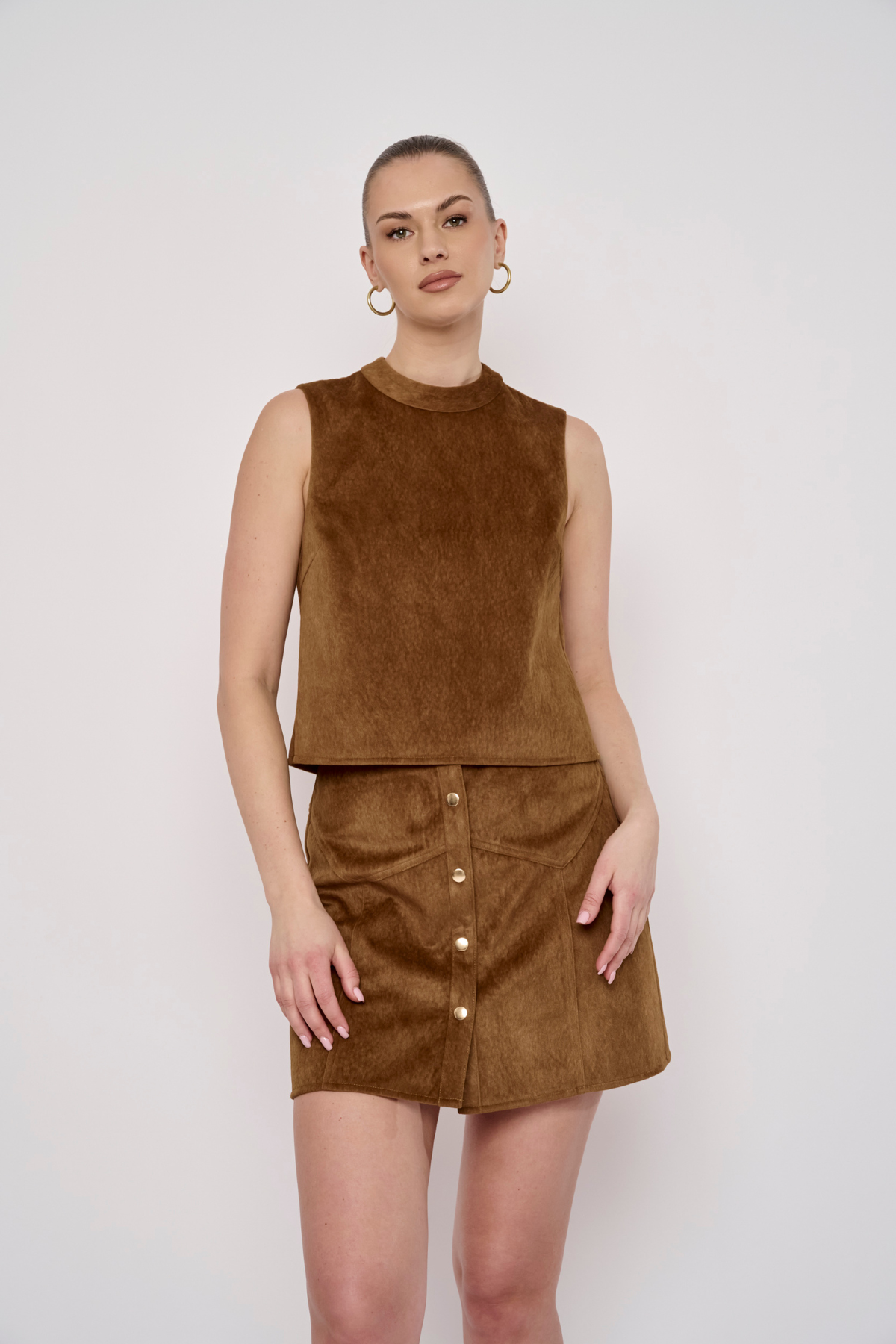 A woman modeling a brown suede-like sleeveless top and matching mini skirt with front buttons, photographed against a plain light gray studio background.