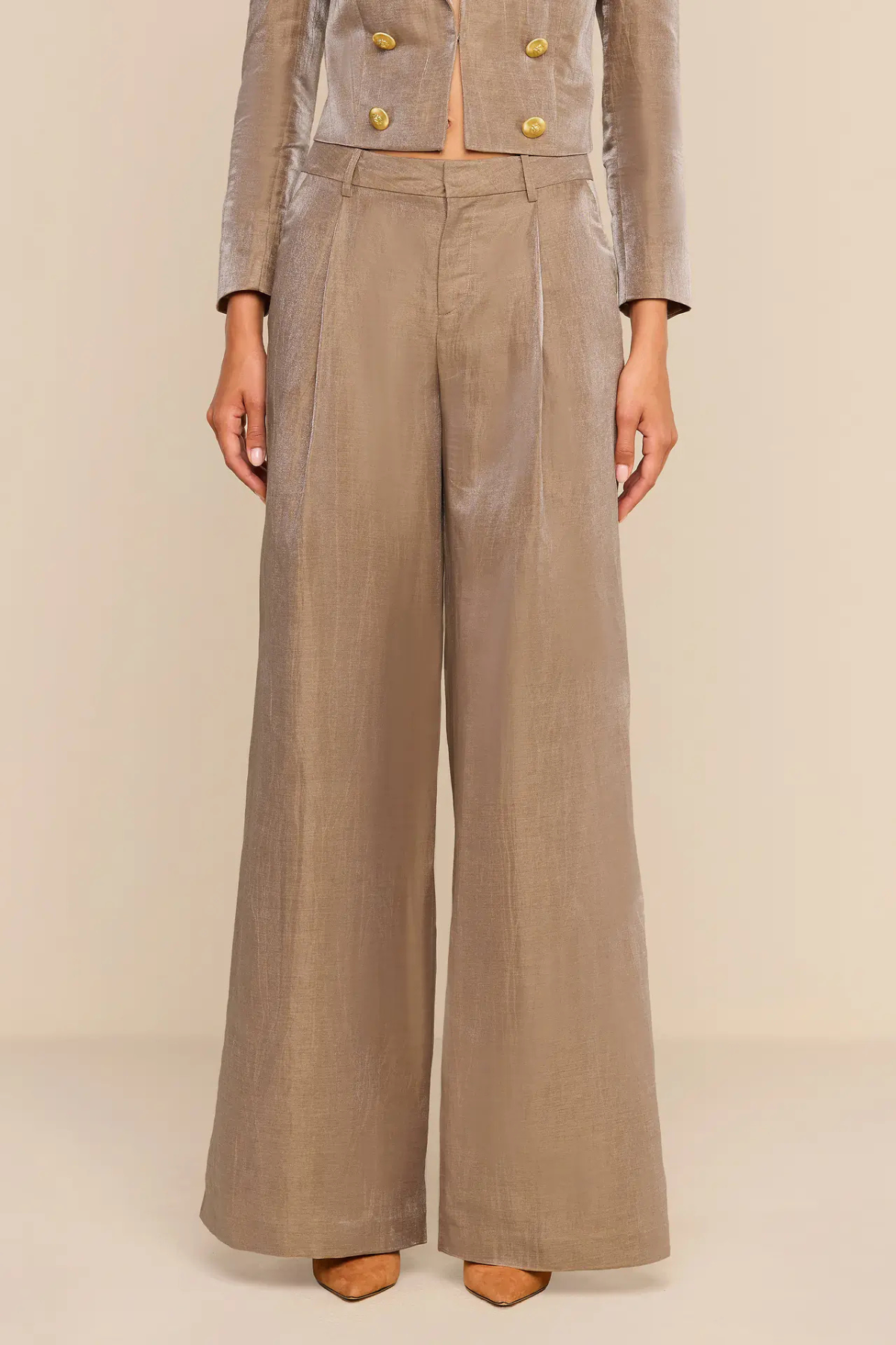 A model wearing a taupe-beige double-breasted wide-leg trouser set with gold buttons, shown from the chest down against a neutral beige background.