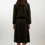 A woman viewed from behind wearing a dark olive green two-piece outfit consisting of a cropped long-sleeve jacket and a matching midi skirt, paired with white heels, against a plain light gray studio background.
