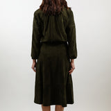 A woman viewed from behind wearing a dark olive green two-piece outfit consisting of a cropped long-sleeve jacket and a matching midi skirt, paired with white heels, against a plain light gray studio background.