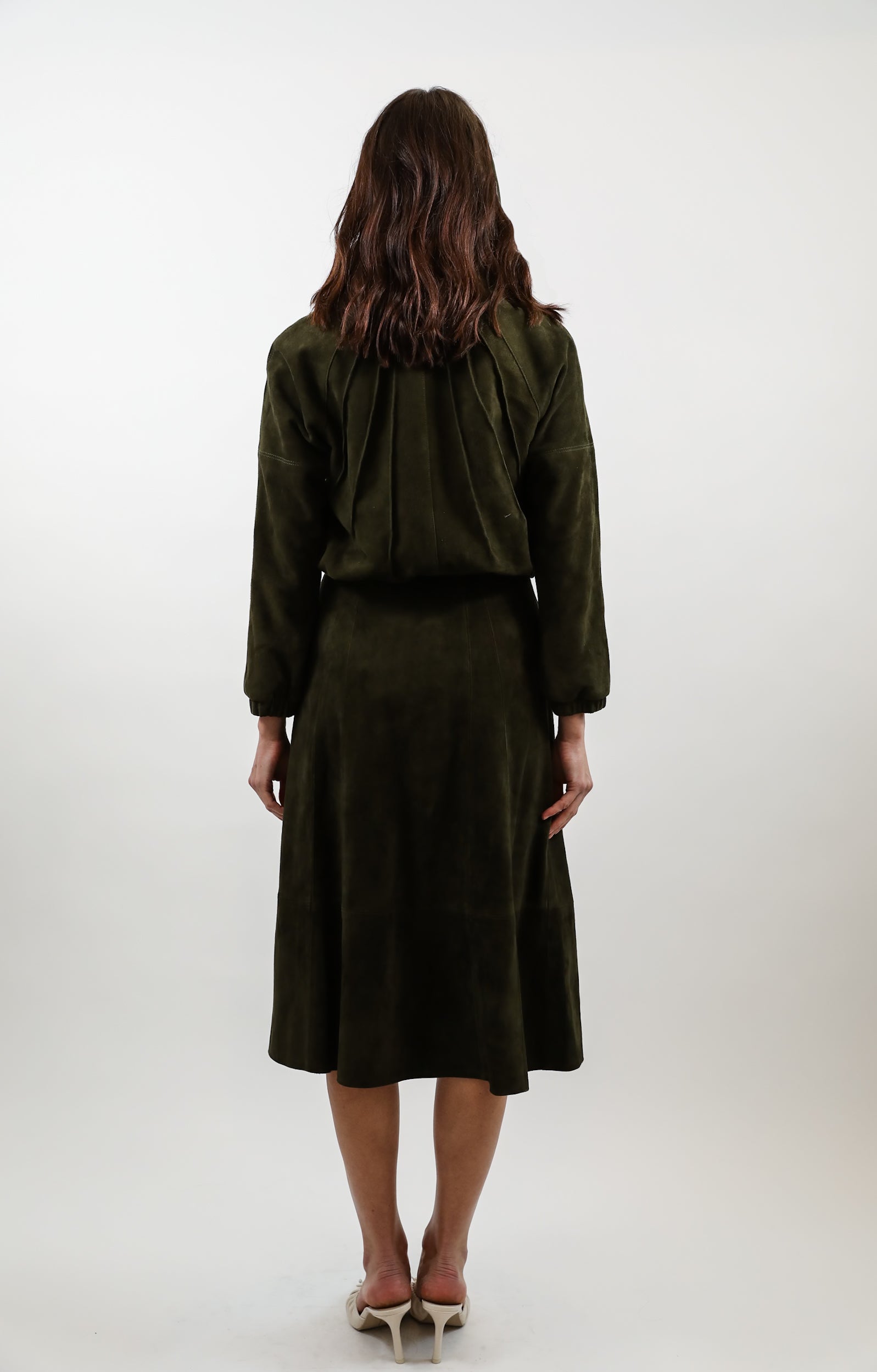 A woman viewed from behind wearing a dark olive green two-piece outfit consisting of a cropped long-sleeve jacket and a matching midi skirt, paired with white heels, against a plain light gray studio background.