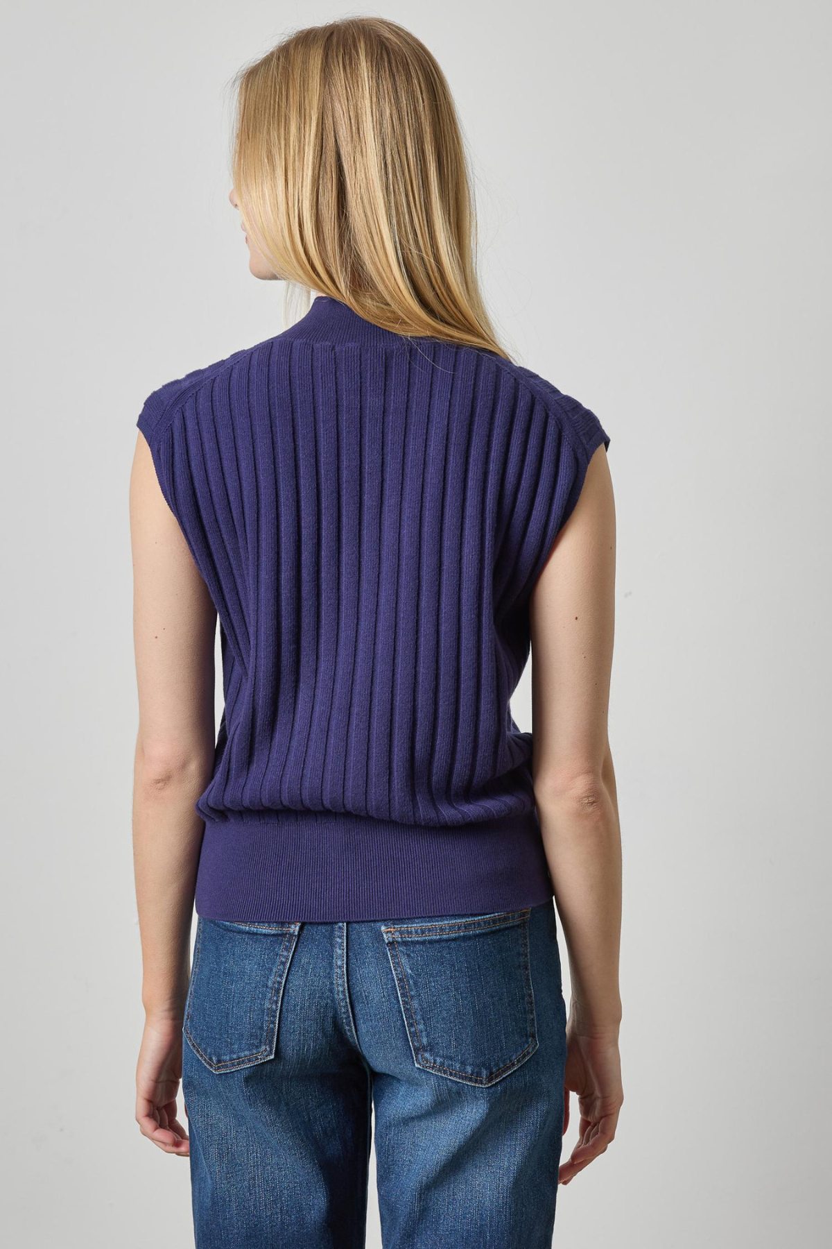 A woman shown from the back wearing a sleeveless, ribbed knit top in a deep purple shade, paired with blue jeans, against a light gray studio background.