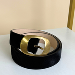 A black velvet belt with a large, square-shaped gold metal buckle, coiled on a white shelf against a beige background.