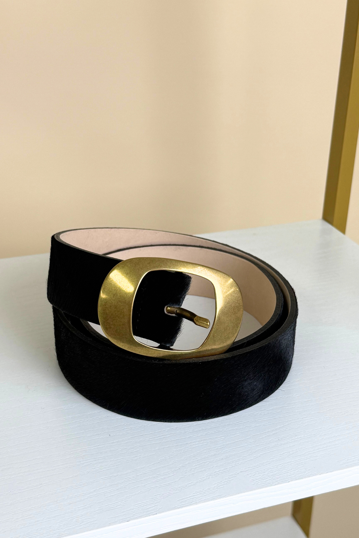A black velvet belt with a large, square-shaped gold metal buckle, coiled on a white shelf against a beige background.
