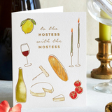 A white greeting card with illustrated food and drink motifs and the text 'to the HOSTESS with the MOSTESS' stands on a tabletop. The card is styled with a wine bottle, candlesticks, flowers, and glassware in the background.