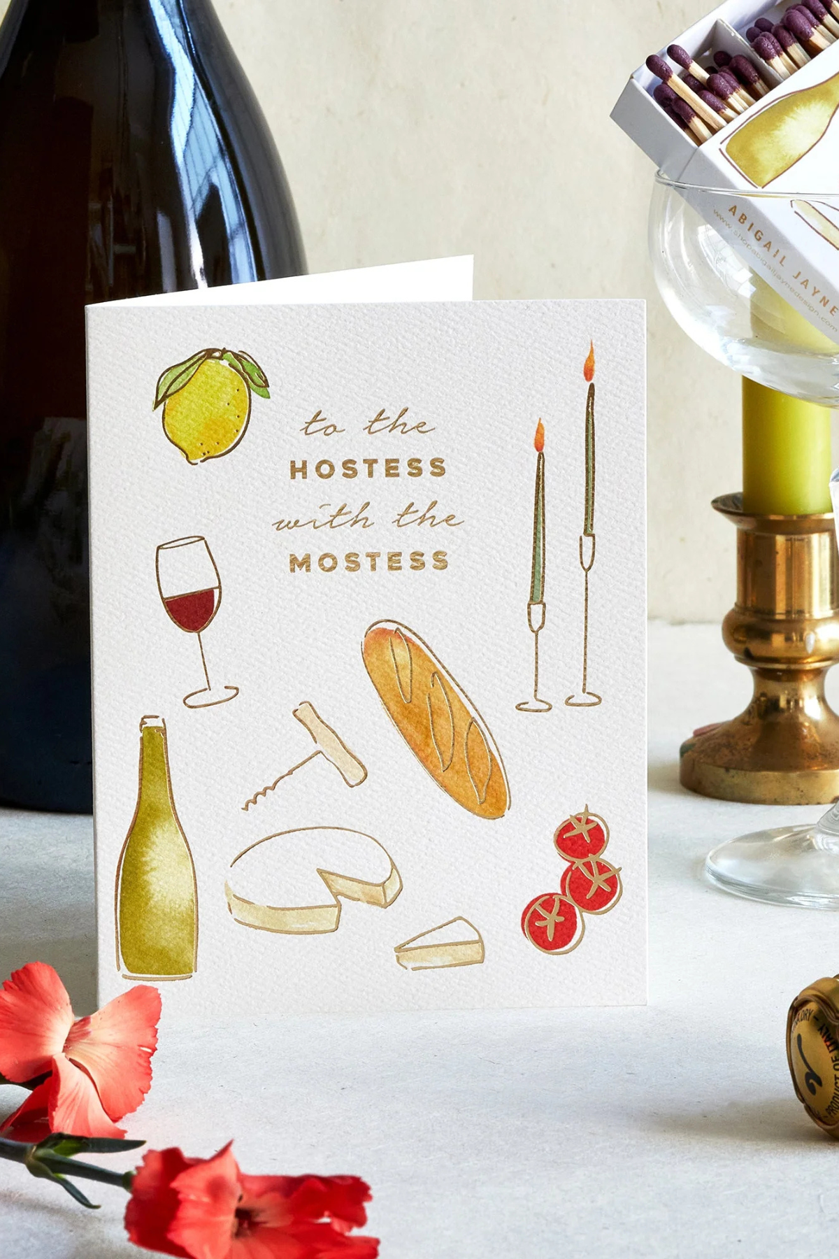 A white greeting card with illustrated food and drink motifs and the text 'to the HOSTESS with the MOSTESS' stands on a tabletop. The card is styled with a wine bottle, candlesticks, flowers, and glassware in the background.