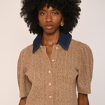 A model with voluminous black curly hair wearing a beige cable-knit cropped short-sleeve top with a dark blue denim collar and gold buttons, paired with dark brown velvet pants.