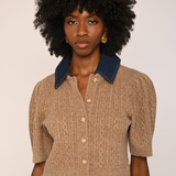 A model with voluminous black curly hair wearing a beige cable-knit cropped short-sleeve top with a dark blue denim collar and gold buttons, paired with dark brown velvet pants.
