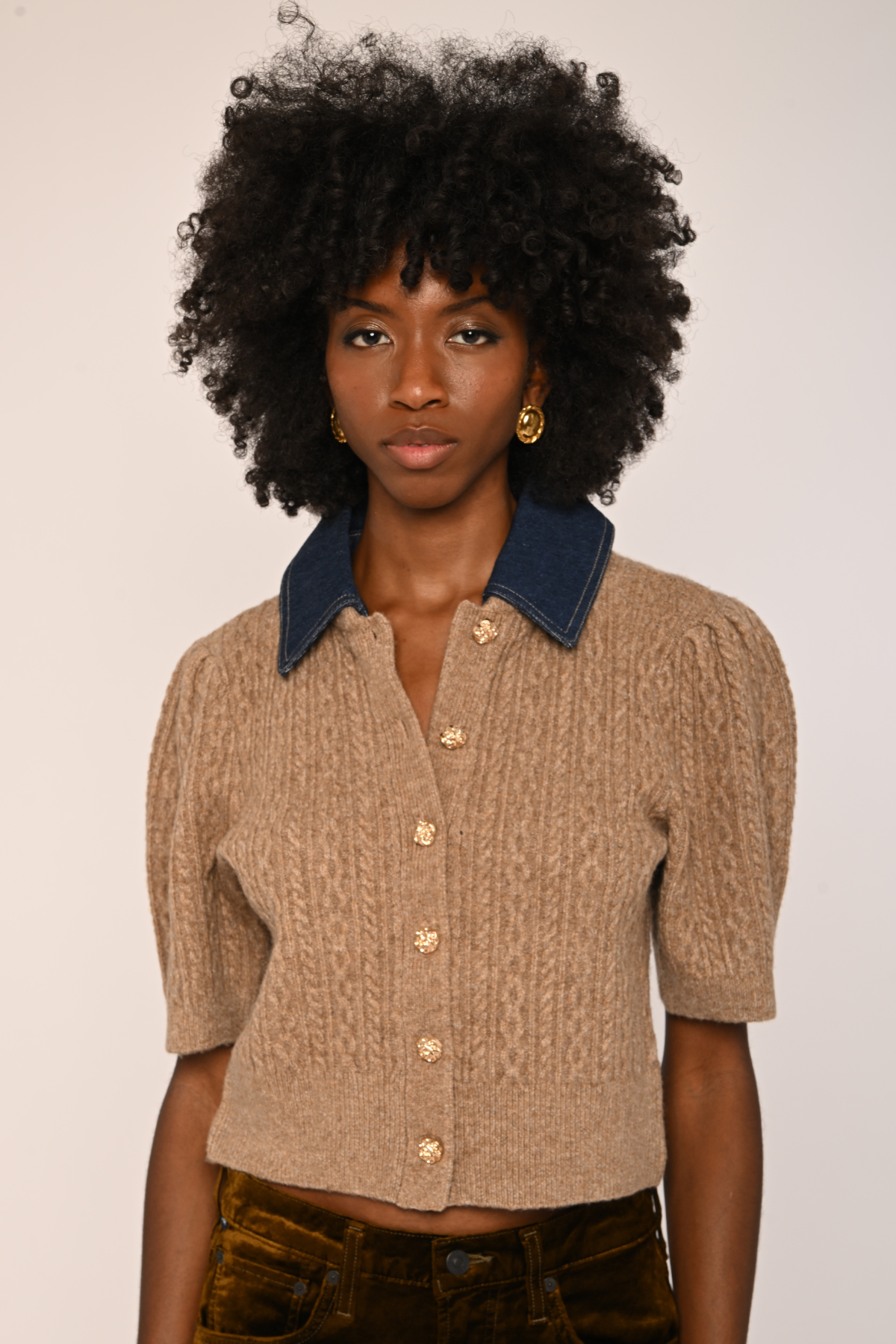 A model with voluminous black curly hair wearing a beige cable-knit cropped short-sleeve top with a dark blue denim collar and gold buttons, paired with dark brown velvet pants.