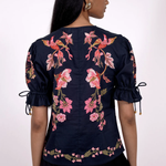 A woman shown from the back wearing a navy blue blouse with pink floral embroidery, puff sleeves with tie details, and a black skirt against a plain white background.