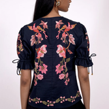 A woman shown from the back wearing a navy blue blouse with pink floral embroidery, puff sleeves with tie details, and a black skirt against a plain white background.