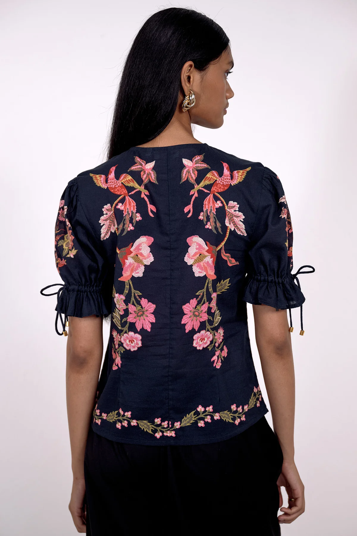 A woman shown from the back wearing a navy blue blouse with pink floral embroidery, puff sleeves with tie details, and a black skirt against a plain white background.