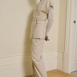 A woman models a light beige two-piece linen set consisting of a sleeveless belted vest and matching wide-leg trousers, layered over a sheer white lace long-sleeve top, in an indoor setting with cream walls and a wooden floor.