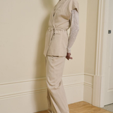A woman models a light beige two-piece linen set consisting of a sleeveless belted vest and matching wide-leg trousers, layered over a sheer white lace long-sleeve top, in an indoor setting with cream walls and a wooden floor.
