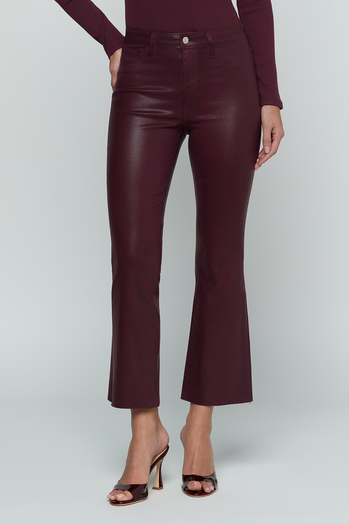 A model wearing high-waisted, cropped flared pants in a deep burgundy faux-leather material, paired with a matching long-sleeve top and dark brown heeled sandals, against a light gray studio background.