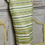 A close-up of a person wearing a fitted midi dress with horizontal stripes in olive green, sage, and cream, standing in front of a distressed gray wooden cabinet.
