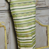 A close-up of a person wearing a fitted midi dress with horizontal stripes in olive green, sage, and cream, standing in front of a distressed gray wooden cabinet.