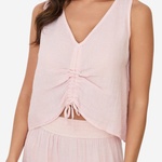 A woman wearing a light pink sleeveless V-neck top with a gathered ruched front and tie detail, paired with a matching skirt against a plain light background.