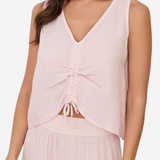 A woman wearing a light pink sleeveless V-neck top with a gathered ruched front and tie detail, paired with a matching skirt against a plain light background.