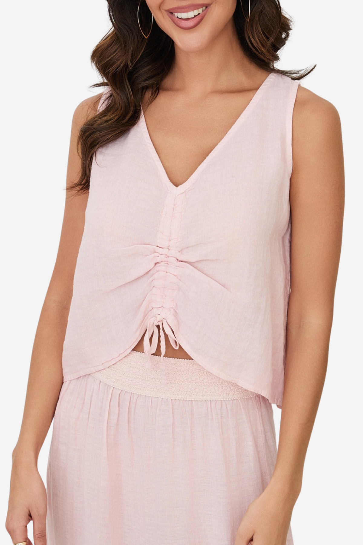 A woman wearing a light pink sleeveless V-neck top with a gathered ruched front and tie detail, paired with a matching skirt against a plain light background.