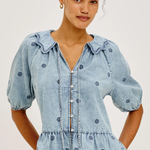 A woman wearing a light blue denim blouse with dark blue embroidered dots, puff sleeves, and a ruffled collar, paired with white pants, against a plain light gray background.