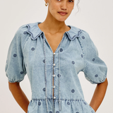 A woman wearing a light blue denim blouse with dark blue embroidered dots, puff sleeves, and a ruffled collar, paired with white pants, against a plain light gray background.