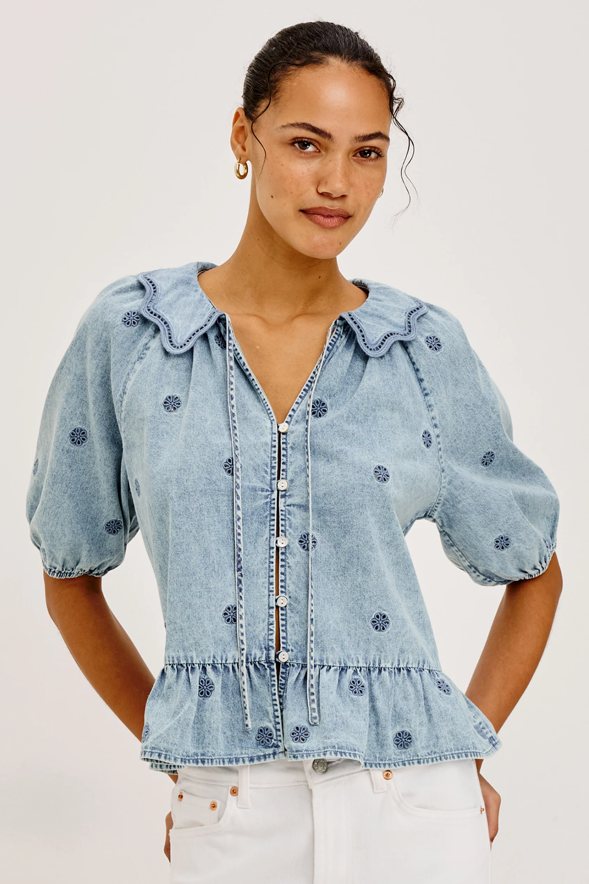 A woman wearing a light blue denim blouse with dark blue embroidered dots, puff sleeves, and a ruffled collar, paired with white pants, against a plain light gray background.