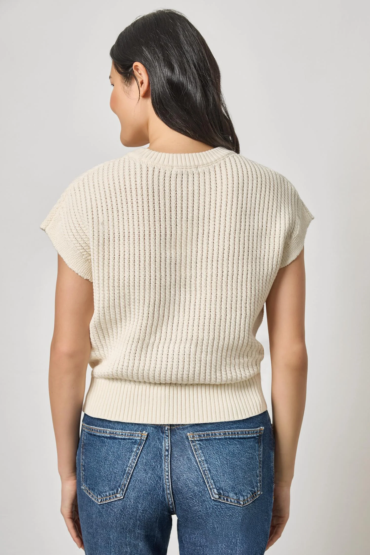 Woman wearing a cream ribbed sweater and blue jeans on a light gray background