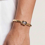 A gold-toned metal bangle bracelet with a rectangular bezel-set dark stone, shown worn on a person's wrist against a light neutral background.