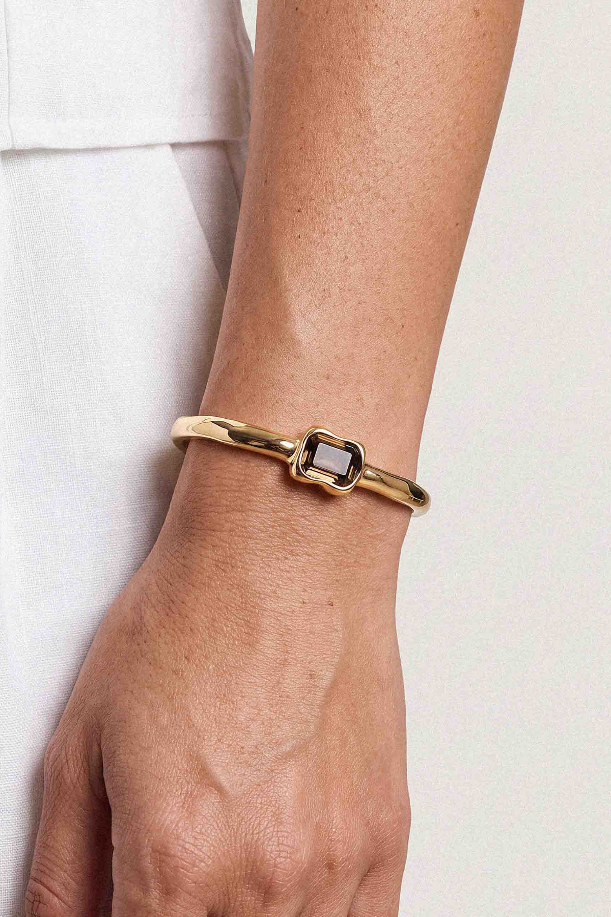 A gold-toned metal bangle bracelet with a rectangular bezel-set dark stone, shown worn on a person's wrist against a light neutral background.