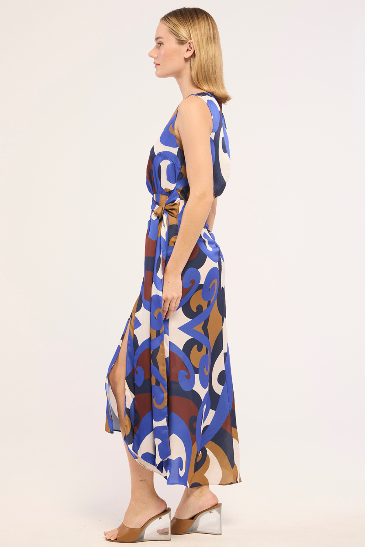A woman modeling a sleeveless midi dress with a blue, brown, and white abstract pattern, shown in profile against a plain light background.