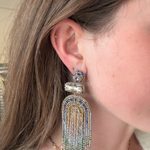 A close-up of a woman's ear wearing a large, ornate statement earring. The earring features a gold-toned metal base adorned with blue and clear rhinestones, with a long fringe tassel made of gold, blue, and clear rhinestones.