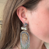 A close-up of a woman's ear wearing a large, ornate statement earring. The earring features a gold-toned metal base adorned with blue and clear rhinestones, with a long fringe tassel made of gold, blue, and clear rhinestones.