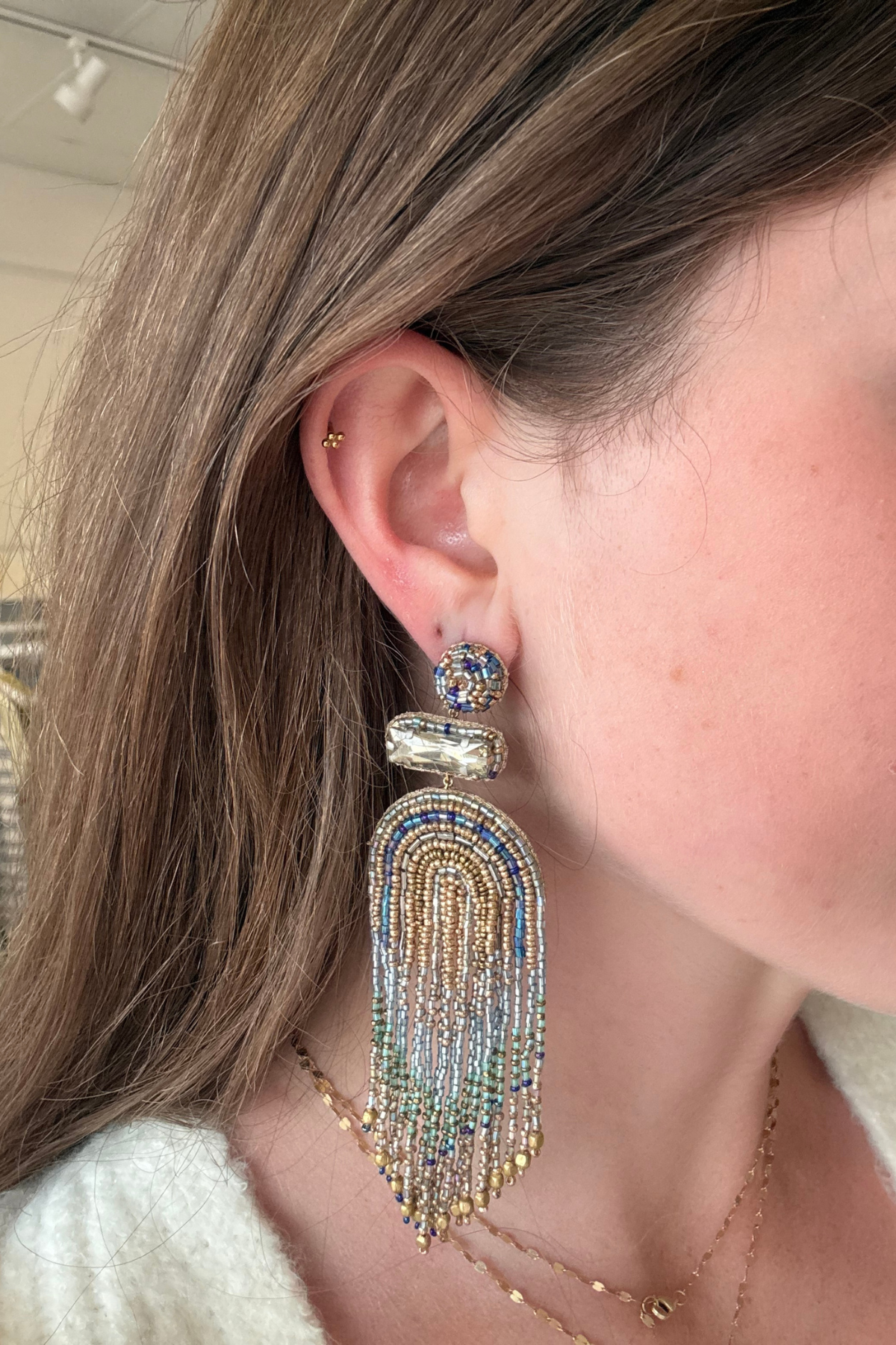 A close-up of a woman's ear wearing a large, ornate statement earring. The earring features a gold-toned metal base adorned with blue and clear rhinestones, with a long fringe tassel made of gold, blue, and clear rhinestones.