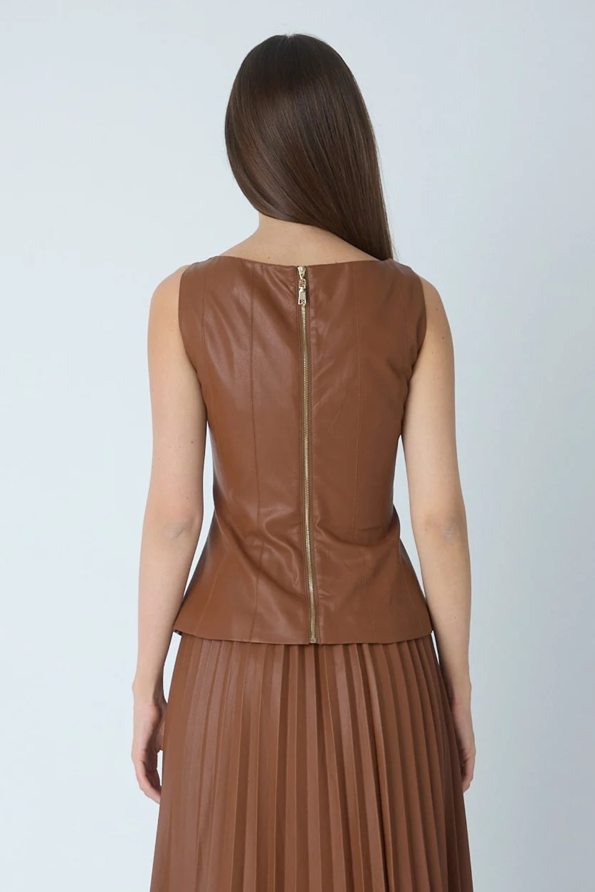 A woman shown from the back wearing a sleeveless brown faux-leather top with a gold-tone zipper and a matching brown pleated skirt, against a light gray studio background.
