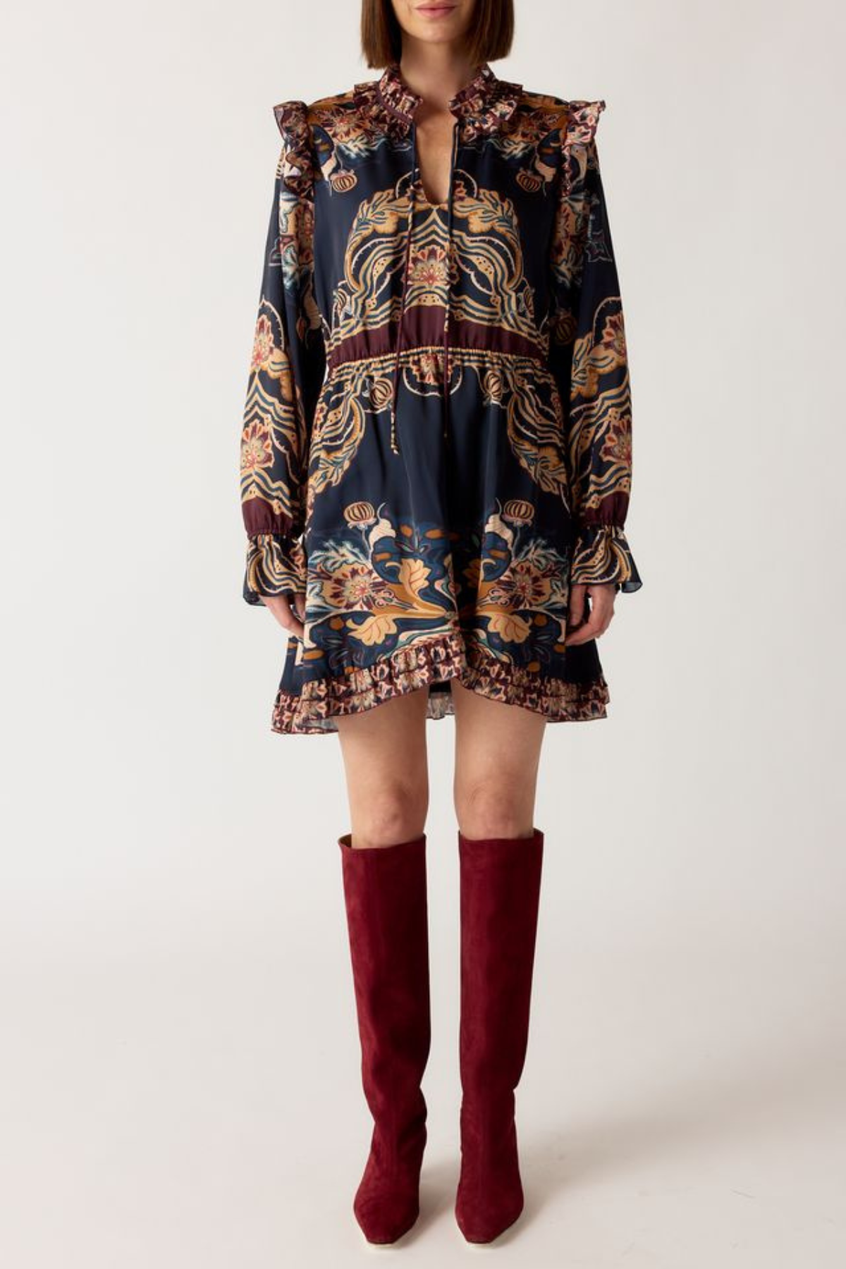 A model wearing a dark navy blue mini dress with a detailed gold, brown, and red patterned print, paired with burgundy over-the-knee boots, against a plain light gray background.