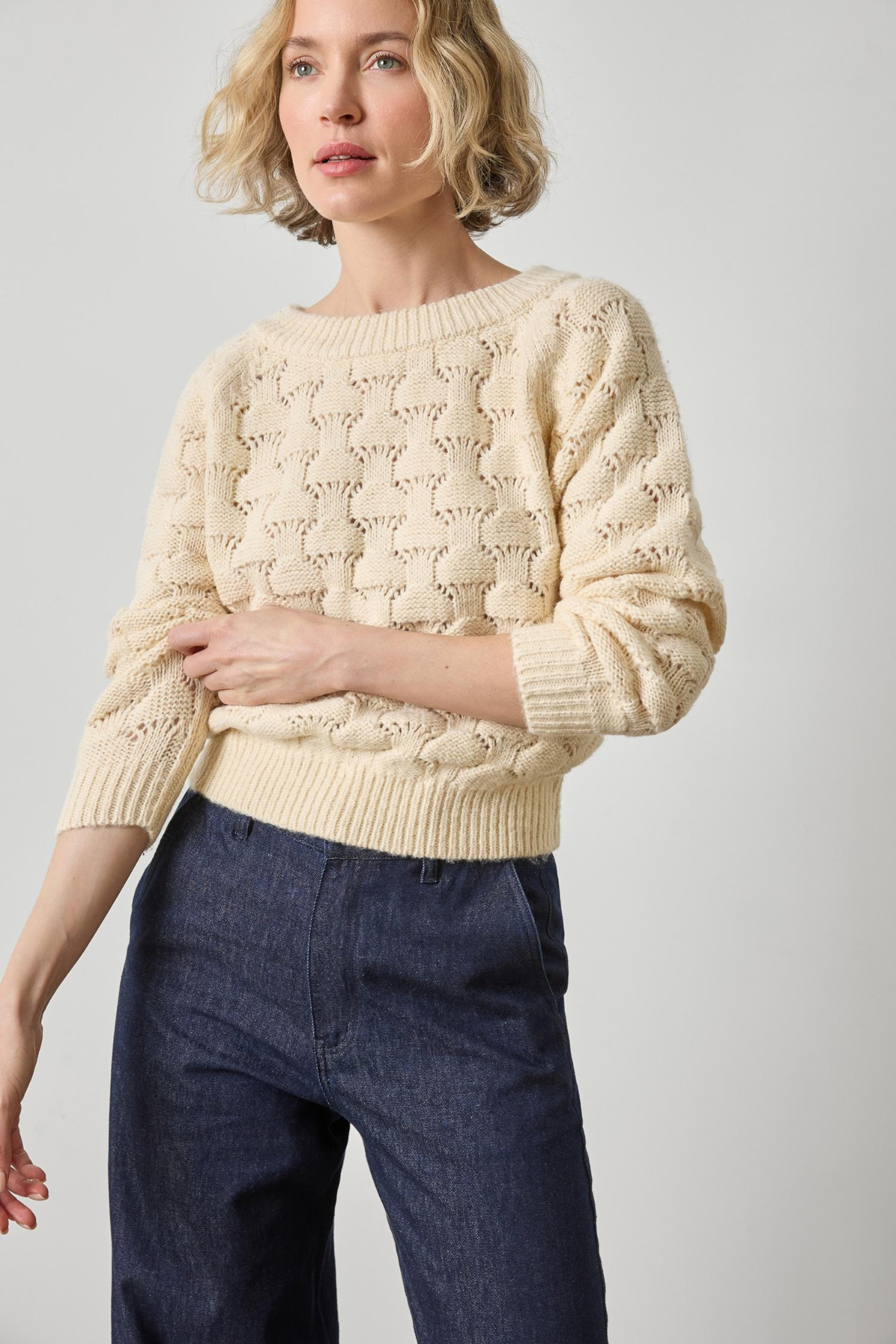 Woman wearing a cream knitted sweater and dark blue jeans on a light gray background