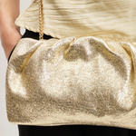 A close-up of a person wearing a shimmering metallic gold clutch bag with a gold chain strap, styled with a cream pleated top and black pants.