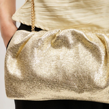 A close-up of a person wearing a shimmering metallic gold clutch bag with a gold chain strap, styled with a cream pleated top and black pants.