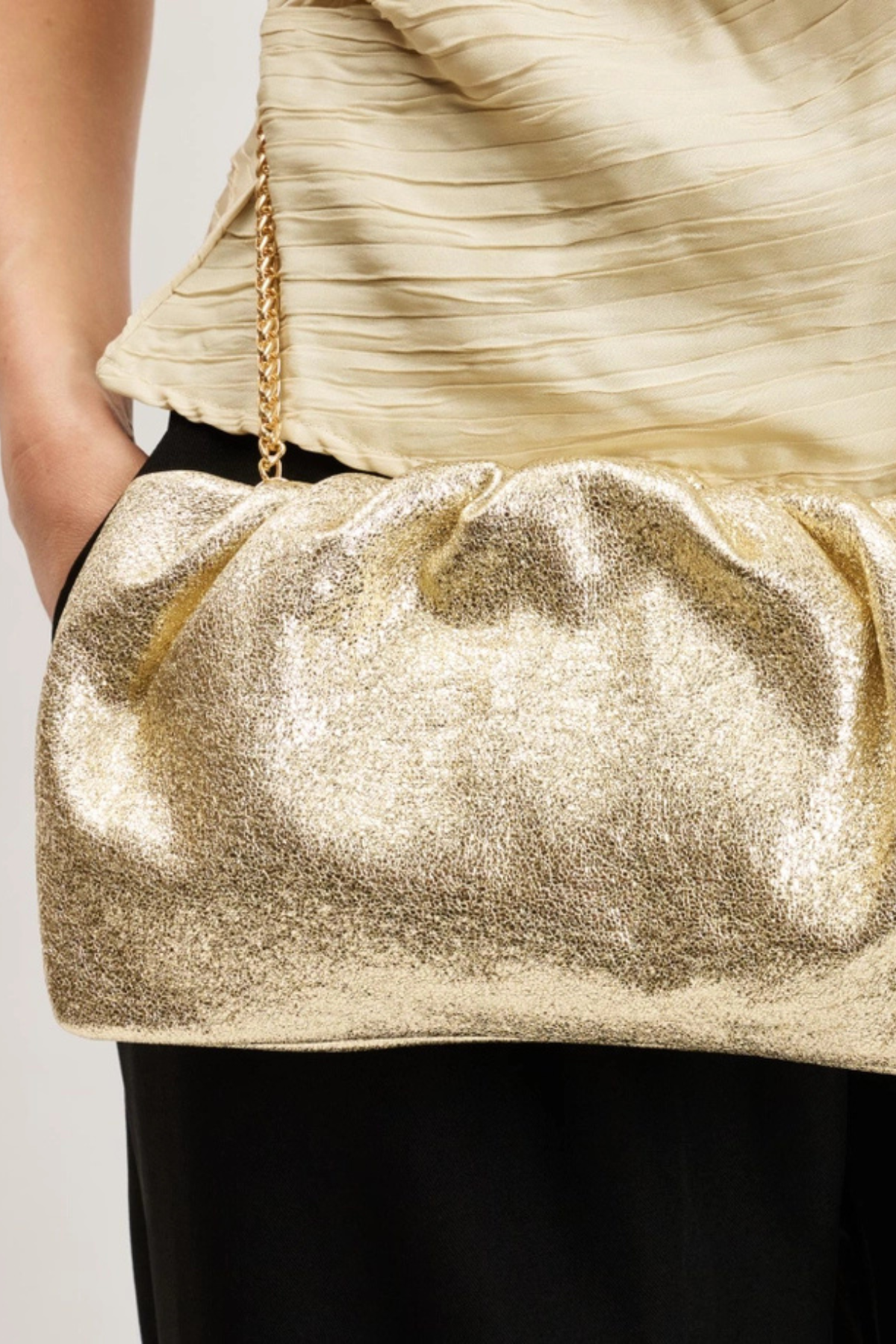 A close-up of a person wearing a shimmering metallic gold clutch bag with a gold chain strap, styled with a cream pleated top and black pants.