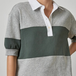 A woman wearing a color-blocked rugby-style polo shirt with heather gray and dark green horizontal panels, a white collar and placket, and rolled sleeves, paired with blue jeans against a light gray studio background.