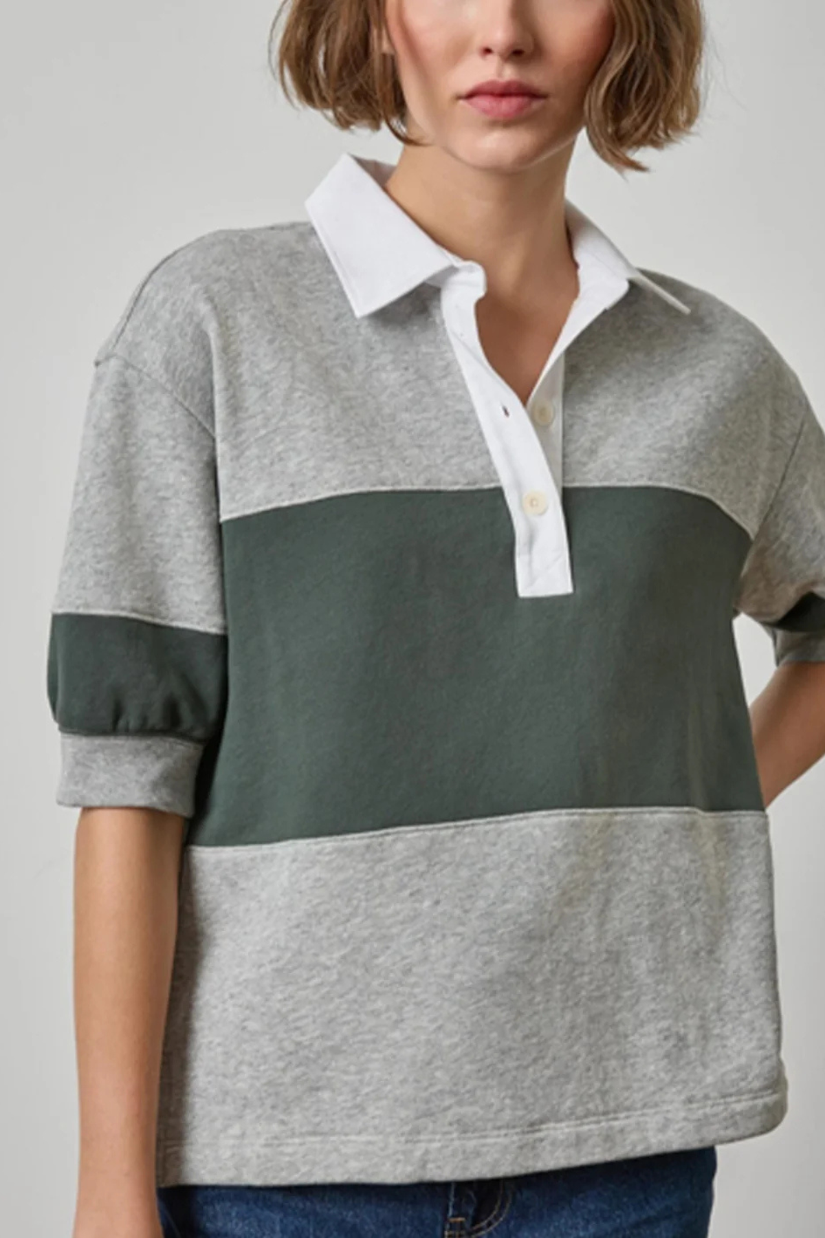 A woman wearing a color-blocked rugby-style polo shirt with heather gray and dark green horizontal panels, a white collar and placket, and rolled sleeves, paired with blue jeans against a light gray studio background.