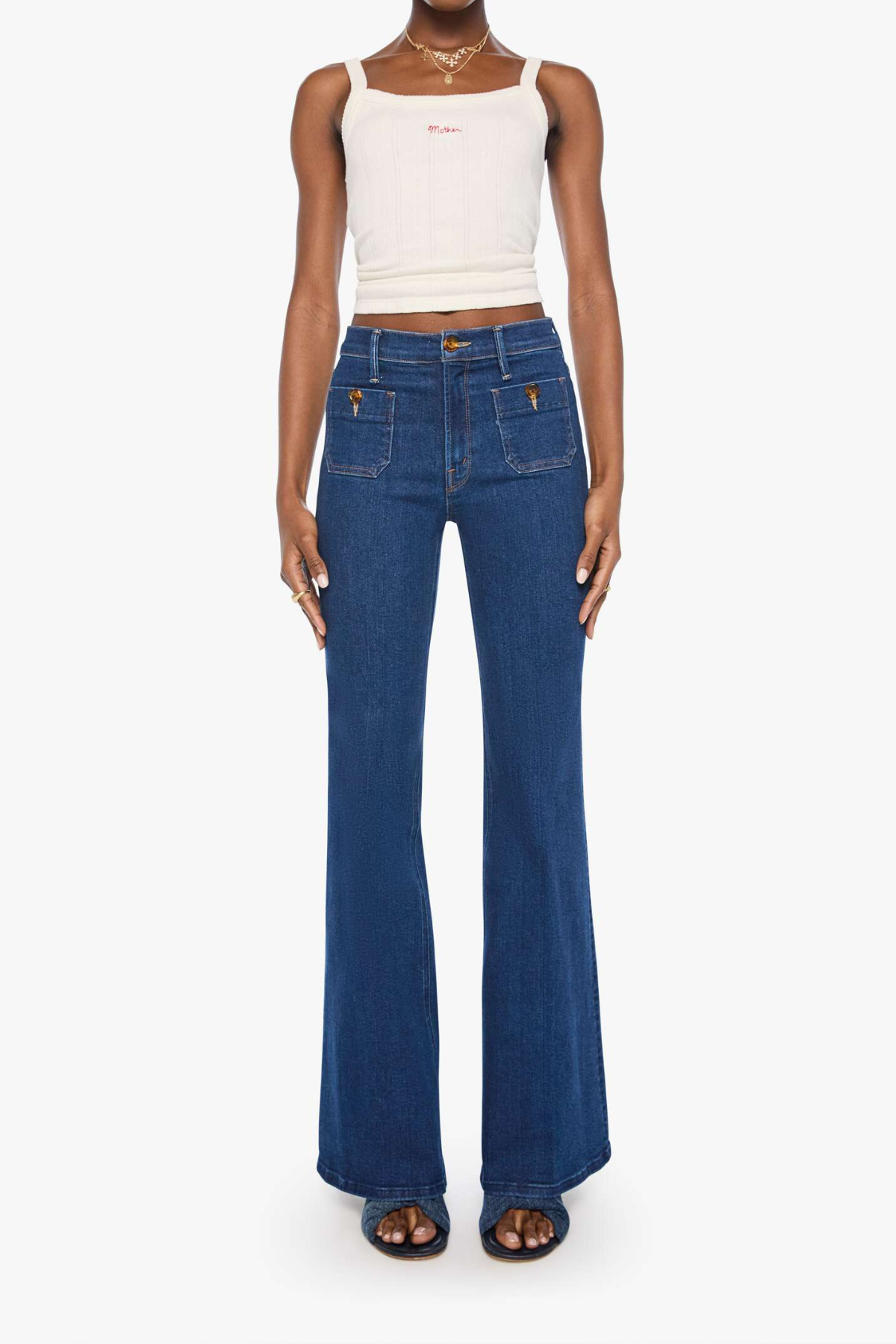 A model wearing high-waisted, wide-leg blue denim jeans with a white ribbed tank top and dark brown sandals, shown against a plain white studio background.