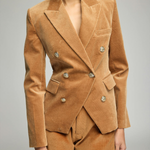 A woman wearing a camel brown velvet double-breasted blazer and matching shorts against a neutral gray studio background.
