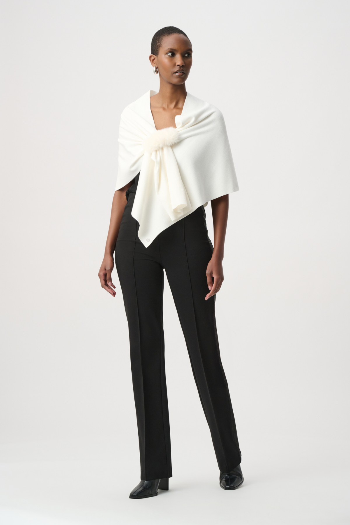A model wearing a cream-colored wrap shawl with a fluffy pom-pom detail at the center, styled over a black top against a light gray studio background.