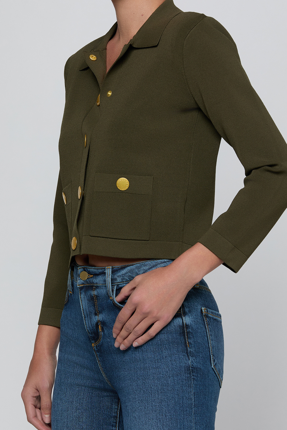 A cropped long-sleeve collared jacket in olive green with gold-tone buttons, worn by a model with blue jeans against a light gray studio background.