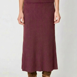 A model wearing a ribbed knit midi skirt in a deep plum purple color, paired with brown suede ankle boots, shown from the waist down against a light gray background.