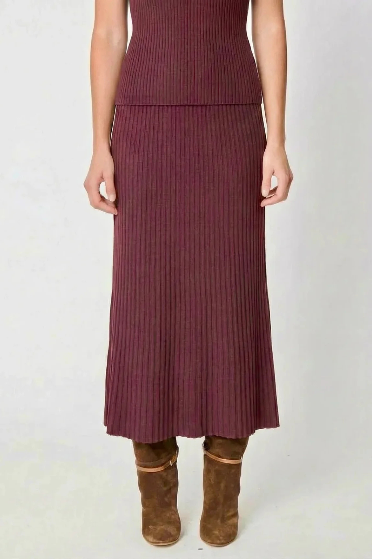 A model wearing a ribbed knit midi skirt in a deep plum purple color, paired with brown suede ankle boots, shown from the waist down against a light gray background.
