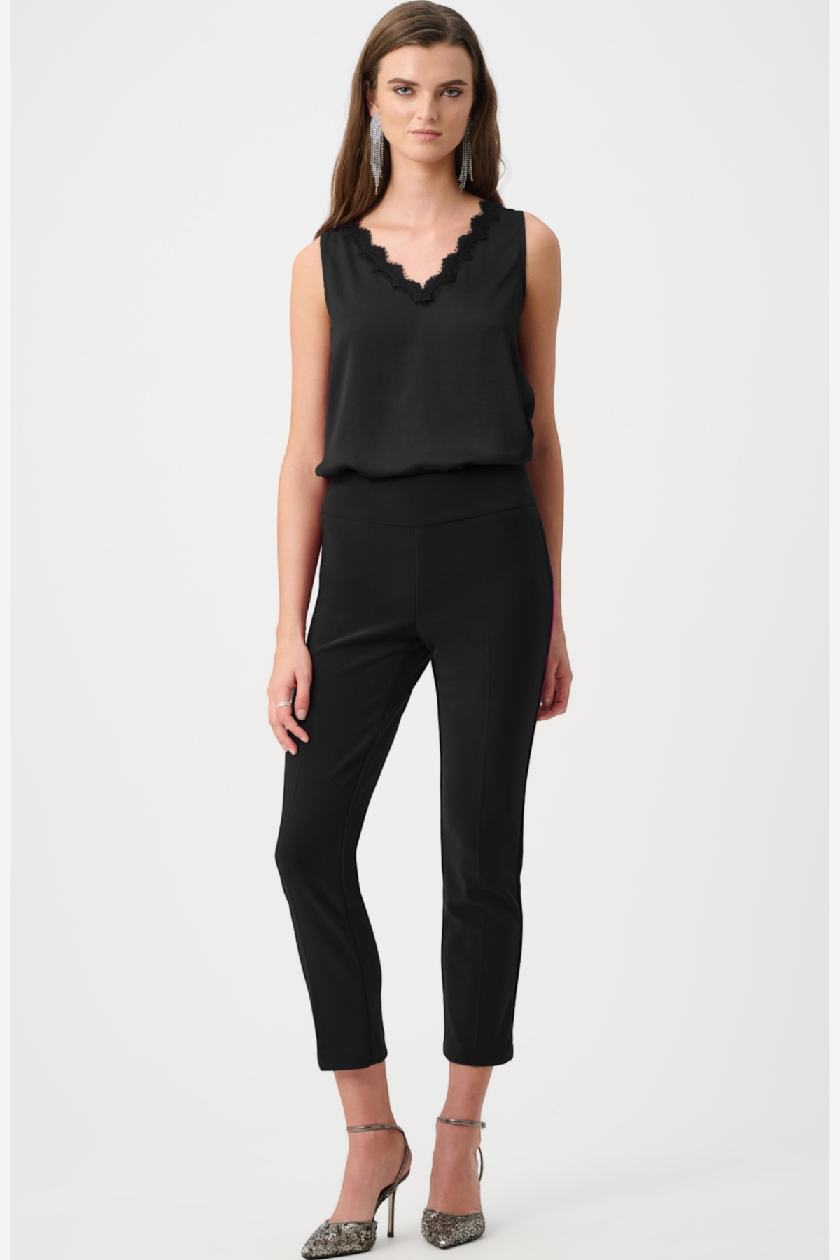A woman modeling a black sleeveless top with a scalloped V-neckline and black cropped pants, paired with pointed-toe heeled sandals, against a plain light gray studio background.