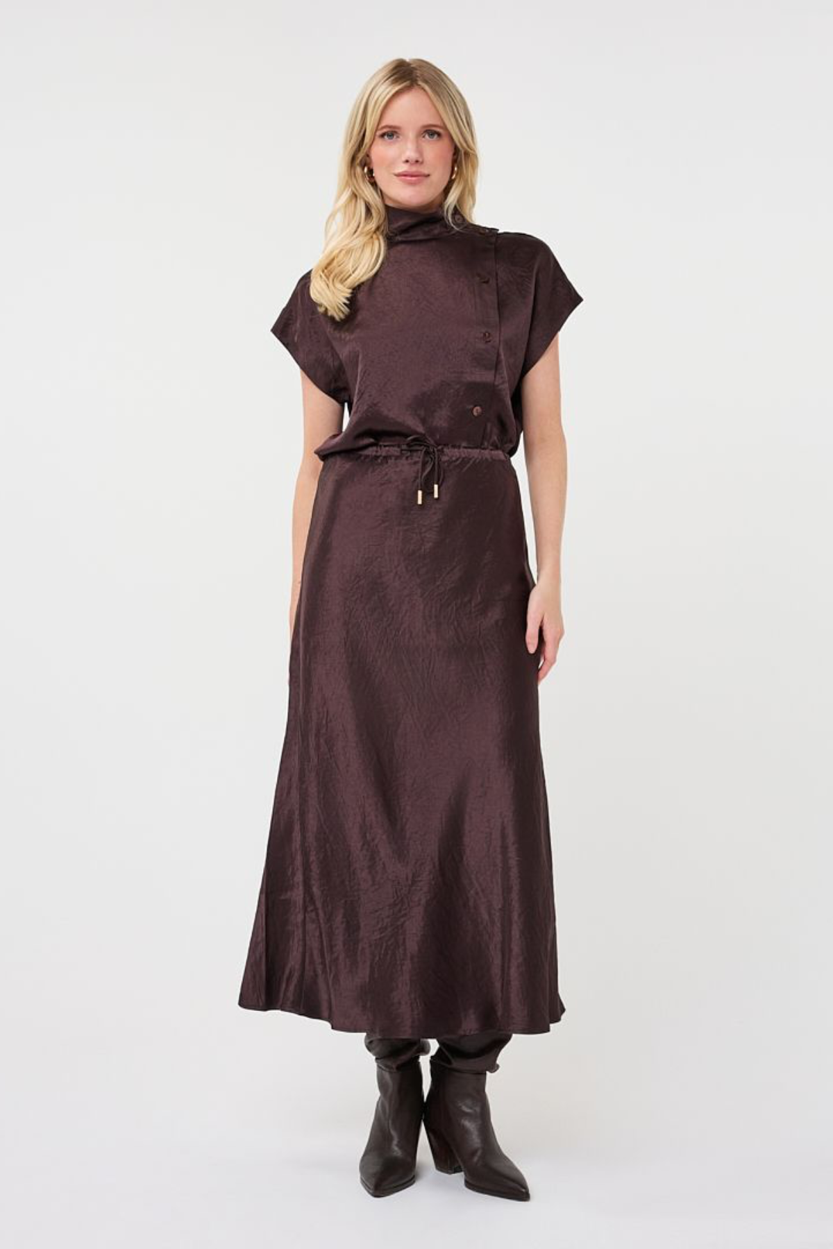 A woman modeling a dark brown short-sleeve midi dress with a high mock neck, drawstring waist, and subtle sheen, paired with black ankle boots against a plain white studio background.